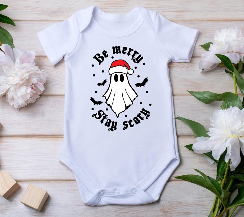 May include: White baby onesie with a black and white graphic of a ghost wearing a Santa hat. The text reads "Be merry. Stay scary."