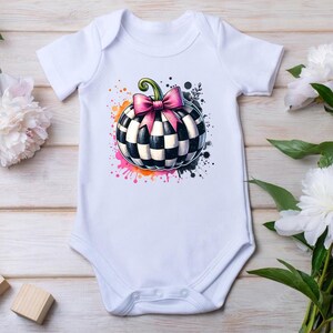May include: White baby onesie featuring a checkered pumpkin design with a pink bow and green stem. The pumpkin is surrounded by colorful paint splatters. The onesie has short sleeves and a snap closure.