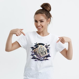 Op de afbeelding: Wit T-shirt met een afbeelding van een halve maan met de tekst "Little Heathen" in zwart en goud. De maan wordt omringd door wolken en vleermuizen.