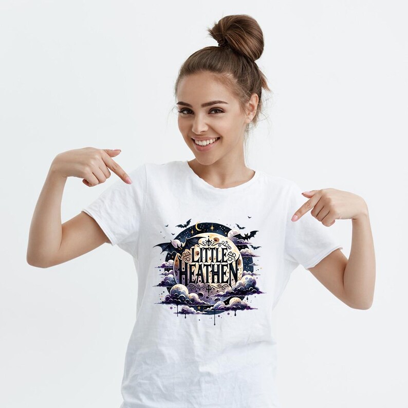 Op de afbeelding: Wit T-shirt met een afbeelding van een halve maan met de tekst "Little Heathen" in zwart en goud. De maan wordt omringd door wolken en vleermuizen.