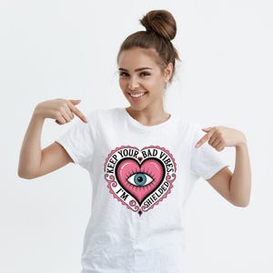 以下が含まれることがあります： 白いTシャツに、ピンクのハートのデザインが施され、目に「Keep Your Bad Vibes I'm Shielded」という文字が書かれています。