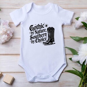 Puede incluir: Body de beb&eacute; blanco con el texto "Gothic by Nature, Southern by Choice" y un gr&aacute;fico de bota de vaquero negra. El dise&ntilde;o est&aacute; en una fuente g&oacute;tica con elementos decorativos. El body est&aacute; hecho de material suave.