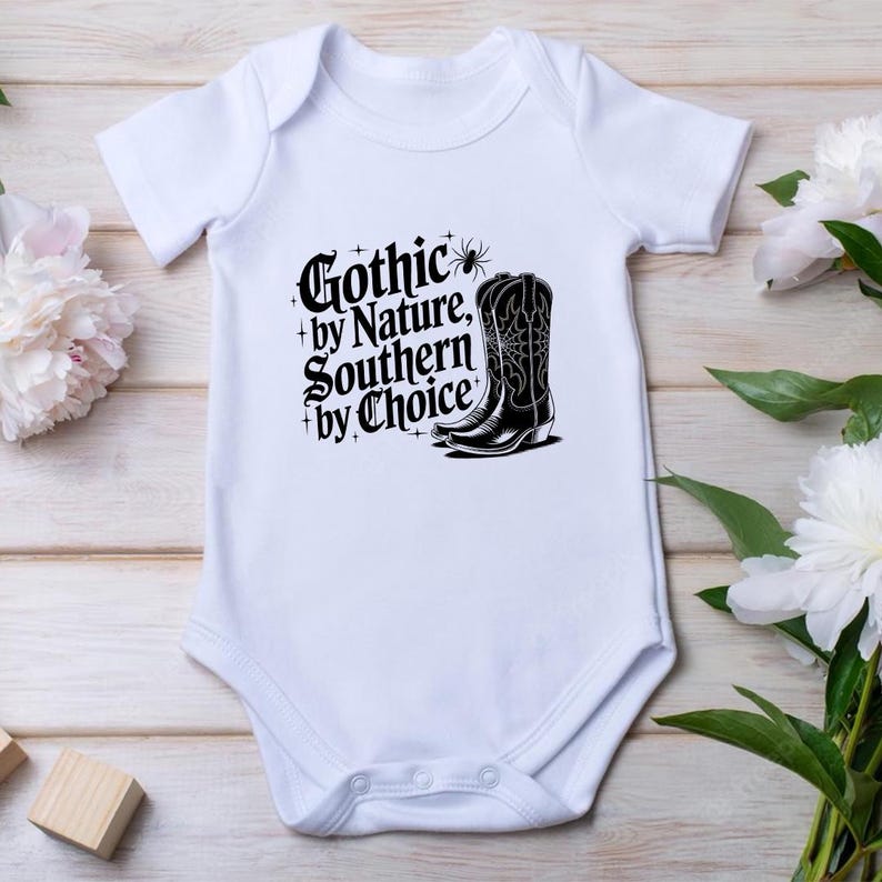 Puede incluir: Body de beb&eacute; blanco con el texto "Gothic by Nature, Southern by Choice" y un gr&aacute;fico de bota de vaquero negra. El dise&ntilde;o est&aacute; en una fuente g&oacute;tica con elementos decorativos. El body est&aacute; hecho de material suave.
