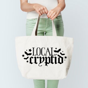 May include: White canvas tote bag with black text that reads "LOCAL cryptid" with bats and stars around the text.