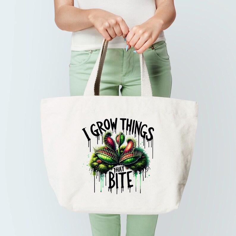 May include: White tote bag with the text "I GROW THINGS THAT BITE" and a colorful illustration of a Venus flytrap. The bag has cream-colored handles and is held by a person.