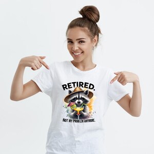 K&ouml;nnte beinhalten: Wei&szlig;es T-Shirt mit einer Illustration eines Waschb&auml;ren, der einen Strohhut tr&auml;gt und einen Cocktail h&auml;lt. Das Design verwendet Aquarelleffekte. Der Text "RETIRED. NOT MY PROBLEM ANYMORE." steht &uuml;ber und unter dem Waschb&auml;ren.