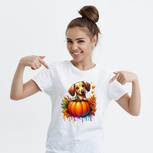 以下が含まれることがあります： 白いTシャツに、漫画のダックスフント犬のデザイン。犬は明るいオレンジ色のカボチャの上から覗いており、秋の葉と水彩絵の具の飛沫に囲まれています。デザインには小さなオレンジ色のハートが含まれています。カラーパレットには、オレンジ、黄色、緑、紫が含まれています。