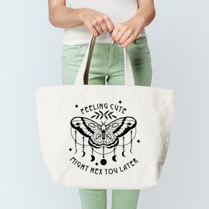 May include: A white tote bag with black text and a graphic of a moth with a crescent moon design. The text reads "Feeling Cute Might Hex You Later".