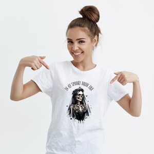 以下が含まれることがあります： 白いTシャツに、ベールと王冠をつけた骸骨の女性のイラストが黒と白で描かれています。イラストの上に「In My Spooky Bride Era」という文字が黒で印刷されています。