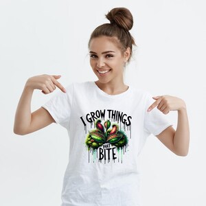 May include: White t-shirt featuring a colorful graphic of a Venus flytrap plant with the text "I GROW THINGS THAT BITE" in black. The design includes green, red, and yellow accents, with dripping paint effects.