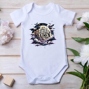 Op de afbeelding: Witte baby romper met een zwart-witte illustratie van een maan met wolken en vleermuizen. De tekst "Little Heathen" staat op de maan.