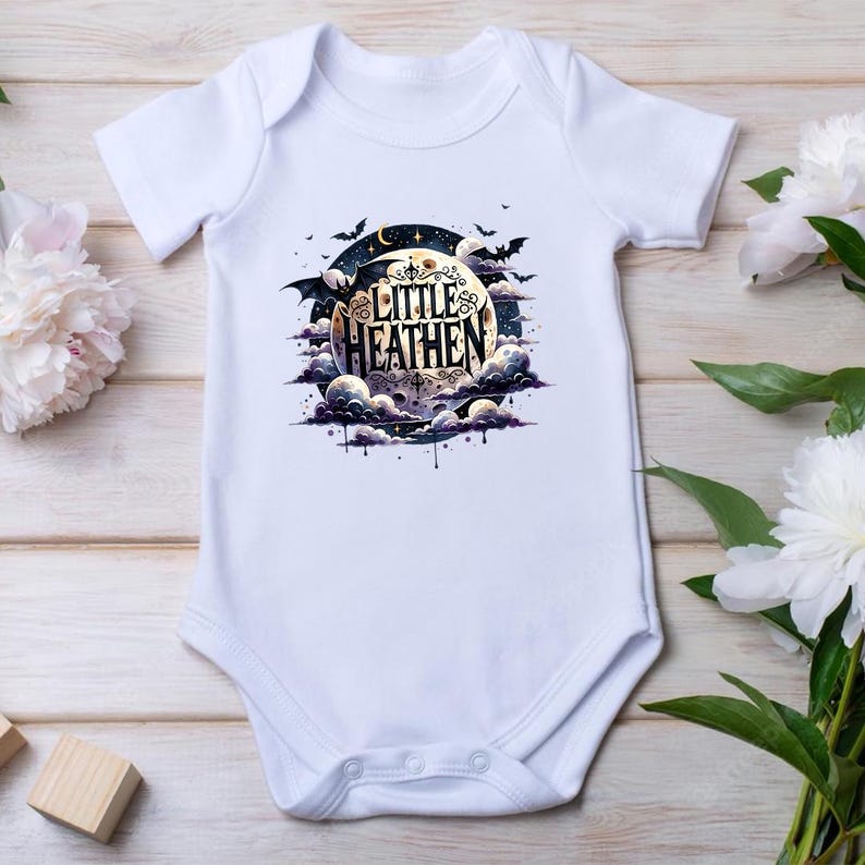 Op de afbeelding: Witte baby romper met een zwart-witte illustratie van een maan met wolken en vleermuizen. De tekst "Little Heathen" staat op de maan.