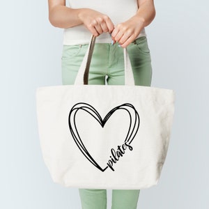 K&ouml;nnte beinhalten: Cremefarbene Tragetasche mit schwarzem Herzdesign und dem Wort "pilates" in Schreibschrift. Die Tasche hat stabile Griffe und wird von einer Person gehalten. Die Tasche besteht aus strapazierf&auml;higem Material, ideal f&uuml;r den Transport von Trainingsger&auml;ten.