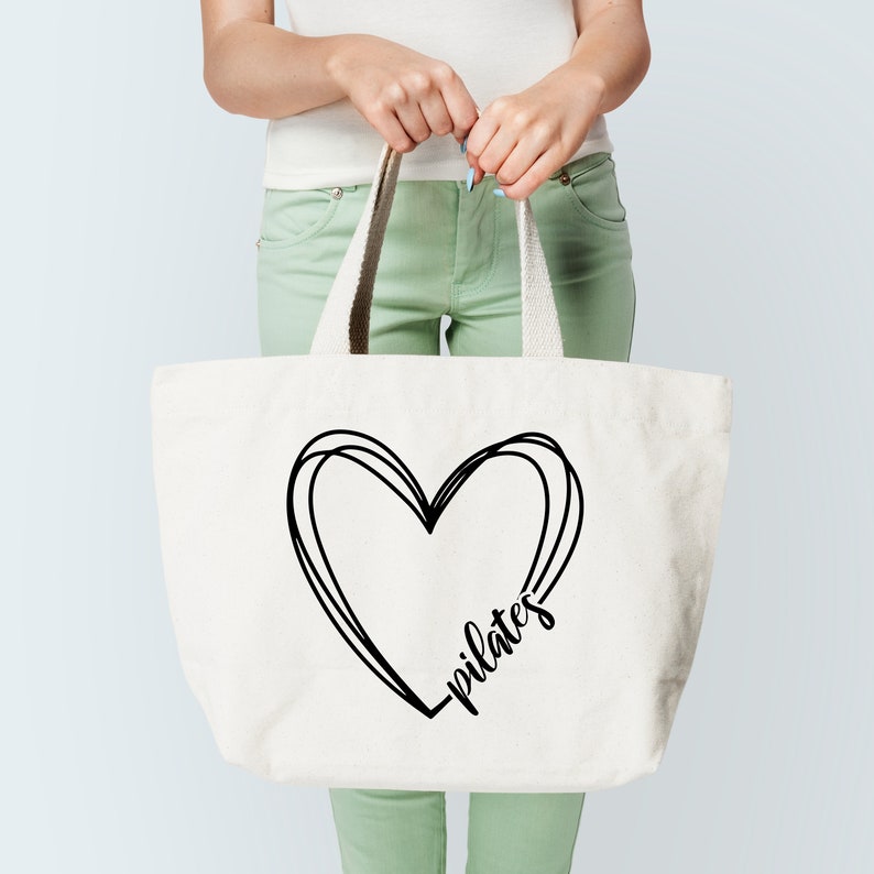 K&ouml;nnte beinhalten: Cremefarbene Tragetasche mit schwarzem Herzdesign und dem Wort "pilates" in Schreibschrift. Die Tasche hat stabile Griffe und wird von einer Person gehalten. Die Tasche besteht aus strapazierf&auml;higem Material, ideal f&uuml;r den Transport von Trainingsger&auml;ten.