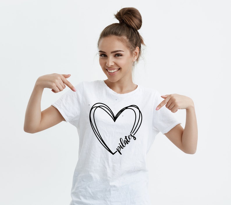 K&ouml;nnte beinhalten: Wei&szlig;es T-Shirt mit einem schwarzen Herzdesign und dem Wort "pilates" in Schreibschrift. Das Herz ist mit drei geschwungenen Linien umrandet. Das Shirt besteht aus einem weichen Material.