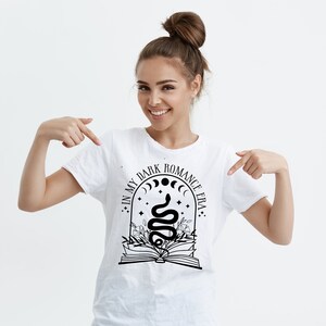 Puede incluir: Camiseta blanca con un dise&ntilde;o gr&aacute;fico negro que presenta una serpiente, un libro y el texto "In My Dark Romance Era."