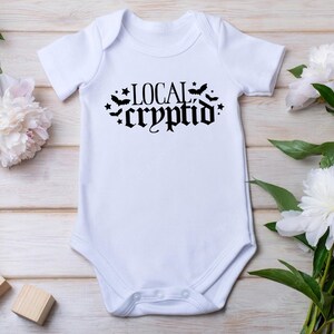 May include: White baby onesie with black text that reads "LOCAL cryptid" with bats and stars around the text.