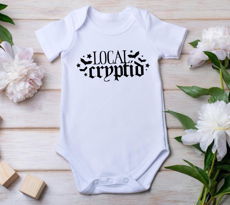 May include: White baby onesie with black text that reads "LOCAL cryptid" with bats and stars around the text.