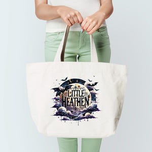 Op de afbeelding: Een witte tote bag met een zwarte en paarse afbeelding van een halve maan met vleermuizen die eromheen vliegen. De tekst "Little Heathen" staat in een gestileerd lettertype op de maan.