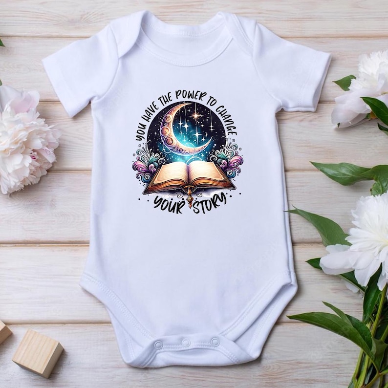 May include: White baby bodysuit with a colorful graphic. The design features an open book with a crescent moon and stars, surrounded by the text "YOU HAVE THE POWER TO CHANGE YOUR STORY."