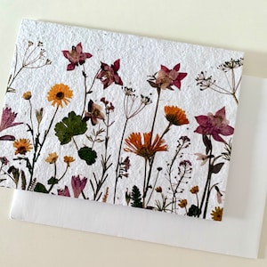 May include: A white card with pressed flowers and leaves. The flowers are in shades of pink, purple, yellow, and orange. The card is made of recycled paper.