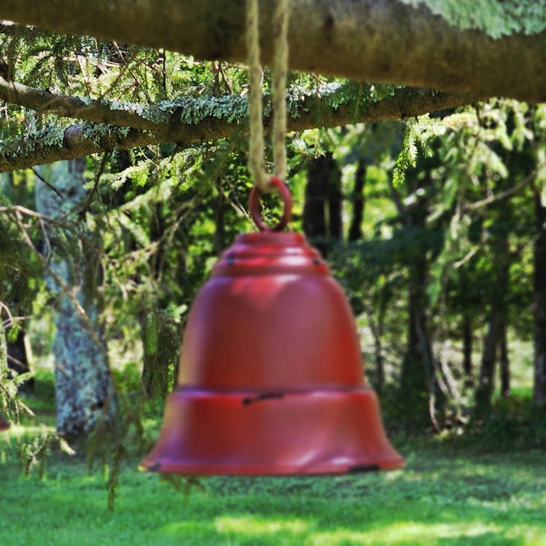 Op de afbeelding: Een rode metalen bel hangend aan een tak met een natuurlijk touw. De bel heeft een klassieke vorm en hangt tegen een achtergrond van groene bomen en gras, wat een buitenomgeving suggereert.