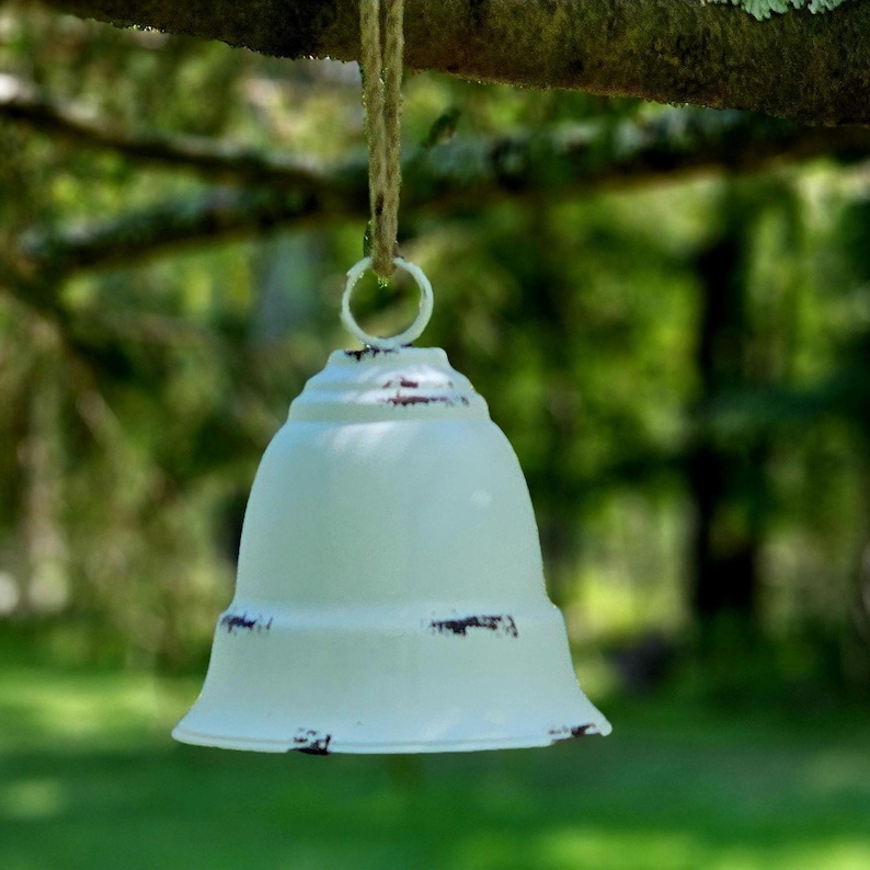 Op de afbeelding: Een verweerde, gebroken witte bel met een rustiek ontwerp hangt aan een natuurlijke touwkoord. De bel heeft een uitlopende basis en een kleine ring aan de bovenkant om op te hangen. De bel hangt aan een tak tegen een vage groene achtergrond.
