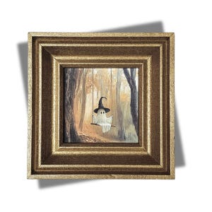 May include: A framed painting depicts a whimsical ghost wearing a witch's hat, swinging on a swing in a forest. The artwork is set within a gold-colored, square frame. The ghost has two small black eyes and is draped in a white sheet.