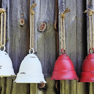 Op de afbeelding: Vier metalen decoratieve bellen, twee wit en twee rood, hangen aan touwen tegen een verweerde houten achtergrond. De bellen hebben een verweerde afwerking, wat een rustieke charme toevoegt.