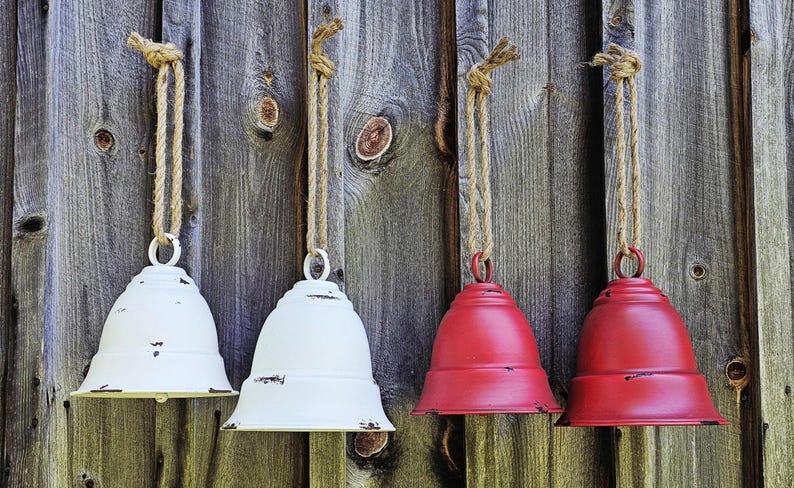 Op de afbeelding: Vier metalen decoratieve bellen, twee wit en twee rood, hangen aan touwen tegen een verweerde houten achtergrond. De bellen hebben een verweerde afwerking, wat een rustieke charme toevoegt.