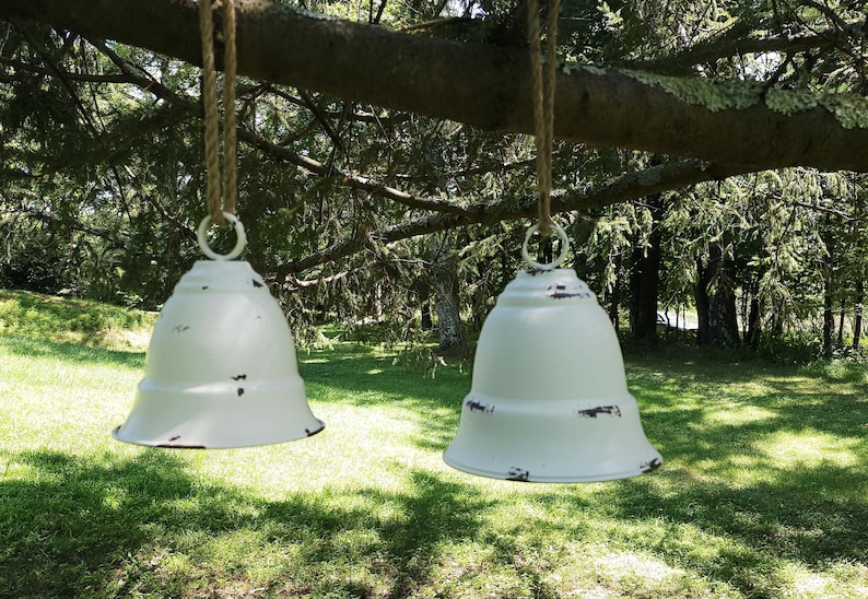 Op de afbeelding: Twee witte, klokvormige hangende plantenbakken met versleten randen hangen aan een tak. De plantenbakken hebben een rustieke, boerderij-esthetiek, ideaal voor buitendecoratie. De achtergrond toont groen gras en bomen.