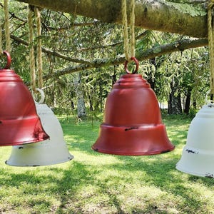 Op de afbeelding: Vier decoratieve metalen bellen, rood en wit, hangend aan een tak. De bellen hebben een verweerde afwerking. De achtergrond bestaat uit groen gras en bomen.