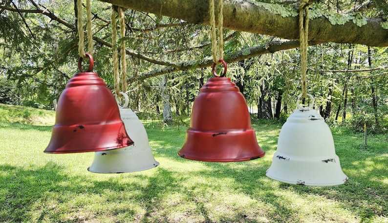 Op de afbeelding: Vier decoratieve metalen bellen, rood en wit, hangend aan een tak. De bellen hebben een verweerde afwerking. De achtergrond bestaat uit groen gras en bomen.