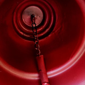 Op de afbeelding: Close-up van een rode bel met een ketting en een rode cilindrische klepel. De binnenkant van de bel heeft concentrische cirkelvormige ribbels. De klepel heeft een kleine, ronde, rode bal aan het einde. De bel is waarschijnlijk van metaal.
