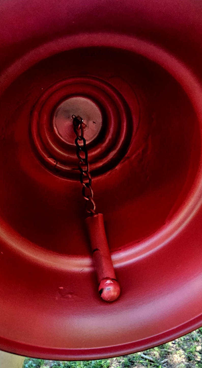 Op de afbeelding: Close-up van een rode bel met een ketting en een rode cilindrische klepel. De binnenkant van de bel heeft concentrische cirkelvormige ribbels. De klepel heeft een kleine, ronde, rode bal aan het einde. De bel is waarschijnlijk van metaal.