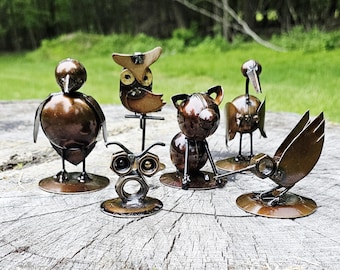 Op de afbeelding: Een verzameling handgemaakte metalen sculpturen van vogels en dieren. De sculpturen omvatten een uil, een vogel, een kat en een kolibrie. De sculpturen zijn gemaakt van metaal met een rustieke bronzen afwerking en staan op een houten oppervlak.