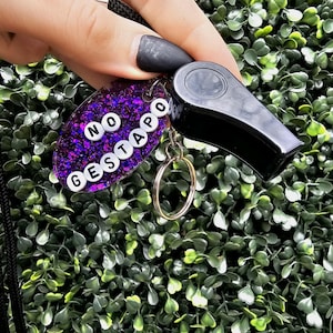 May include: A black whistle with a gold keyring attached to a purple glittery oval keychain. The keychain has the words "NO GESTAPO" spelled out in white letter beads. A black lanyard is attached to the whistle.