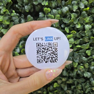 Op de afbeelding: Een ronde witte button met de zwarte tekst LET'S LINK UP!, waarbij het woord LINK in een blauw kader is gemarkeerd, geplaatst boven een vierkante zwart-witte QR-code. De button heeft een gladde, glanzende afwerking en een strak ontwerp.