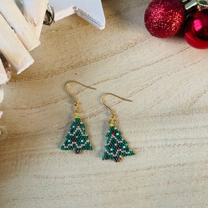 Könnte beinhalten: Ein Paar Ohrringe in Form von Weihnachtsbäumen. Jeder Ohrring ist ein grüner, mit Perlen besetzter Weihnachtsbaum mit roten, weißen und rosa Akzenten. Die Ohrringe haben goldfarbene Haken und werden auf einer hellen Holzoberfläche mit Weihnachtsornamenten im Hintergrund präsentiert.
