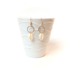 Peut inclure: Une paire de boucles d'oreilles dorées avec un petit pendentif en coquillage de couleur crème. Les boucles d'oreilles sont présentées sur un fond blanc texturé.