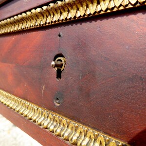 May include: Close-up of a wooden surface with a gold trim and a keyhole. The surface is a dark reddish brown color and the trim is a shiny gold.