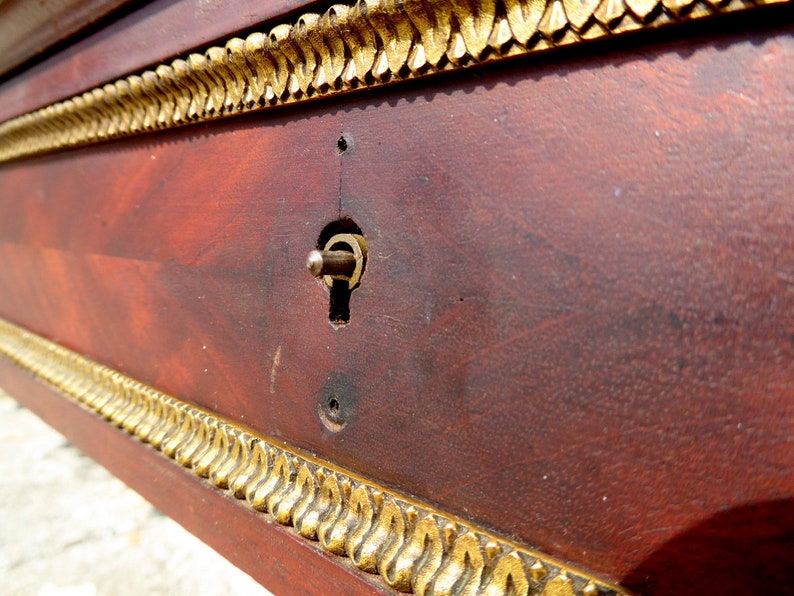 May include: Close-up of a wooden surface with a gold trim and a keyhole. The surface is a dark reddish brown color and the trim is a shiny gold.
