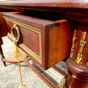 May include: A mahogany console table with gold accents and two drawers. The table has a rope pull on the bottom drawer. The table is in the French Empire style.