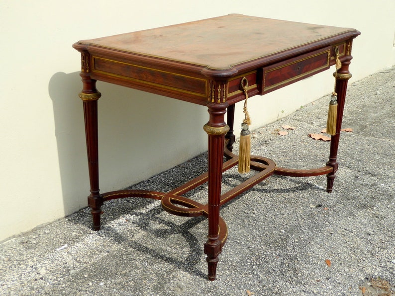May include: A mahogany writing desk with a leather top, a single drawer, and gold accents. The desk has a curved, open base with a decorative design.