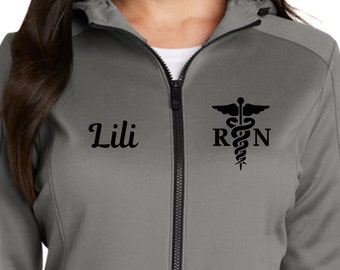 May include: A gray zip-up hoodie with black accents, featuring the name "Lili" and a Registered Nurse (RN) symbol. The jacket has a hood, two front pockets, and thumbhole cuffs. A New Era logo is visible on the sleeve.