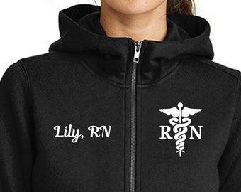 May include: Black zip-up hoodie with a white medical symbol and the text "RN" and "Lily, RN" on the front. The hoodie has a New Era logo on the sleeve.