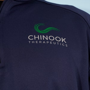 以下が含まれることがあります： 「CHINOOK THERAPEUTICS」と書いてある緑色のロゴが付いたネイビーブルーのクォータージッププルオーバーです。