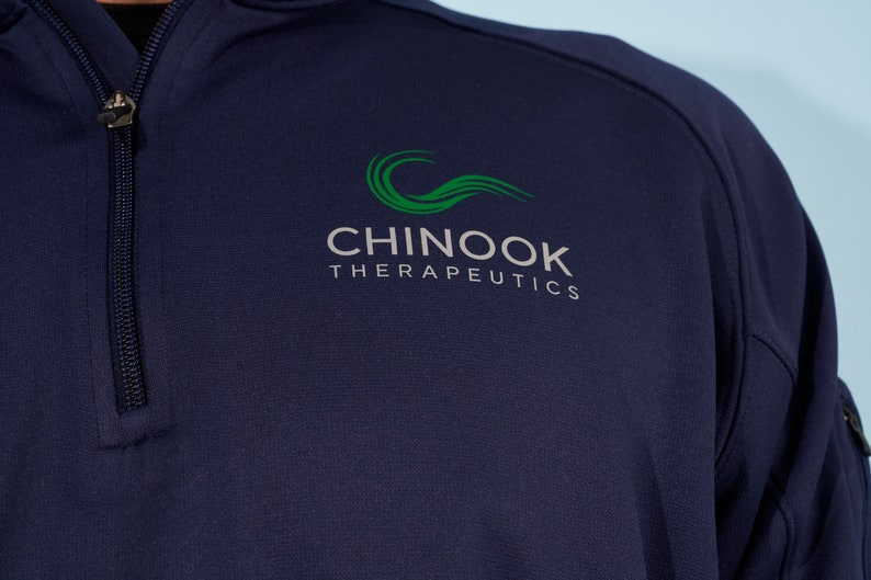 以下が含まれることがあります： 「CHINOOK THERAPEUTICS」と書いてある緑色のロゴが付いたネイビーブルーのクォータージッププルオーバーです。