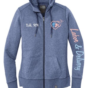 May include: A blue heather zip-up hoodie with a white embroidered heart with baby footprints and a stethoscope. The text "Lili, RN" is embroidered on the left chest and "Labor & Delivery" is embroidered on the right sleeve.