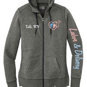 May include: A graphite grey zip-up hoodie with a black zipper and black drawstrings. The hoodie has a heart with baby footprints and a stethoscope on the front and the text "Lili, RN" below it. The left sleeve has the text "Labor & Delivery" in blue.